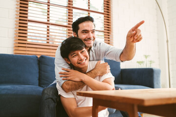Fototapeta premium Happy caucasian and Asian LGBT couple, one sitting on the sofa embracing the other sitting on the floor from the back at home. Diversity of LGBT relationships. A gay couple concept.