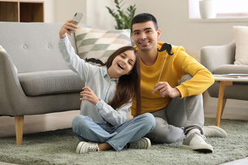 Fototapeta premium Portrait of father and his little daughter with paper mustache taking selfie at home