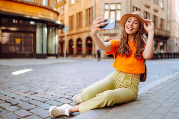 Happy woman holding mobile phone takes selfie using smartphone camera. A tourist walks the streets and enjoys the architecture of the city, taking pictures of the sights. Selfie time. 