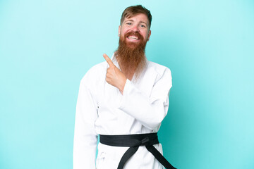 Redhead man with long beard doing karate isolated on blue background pointing to the side to present a product