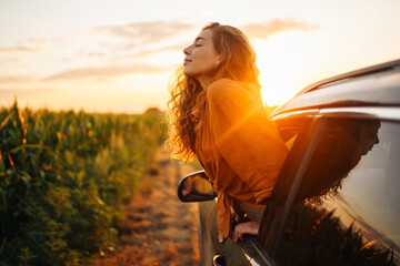 Young happy woman leaning out of the car window enjoying the sunset. The concept of active lifestyle, travel, tourism, nature. - obrazy, fototapety, plakaty