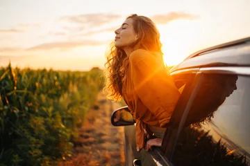 Vlies Fototapete Schokoladenbraun Young happy woman leaning out of the car window enjoying the sunset. The concept of active lifestyle, travel, tourism, nature.