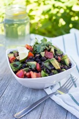 Bowl of healthy salad with lime, avocados, tomatoes, and beans with lemonade water on the gray table