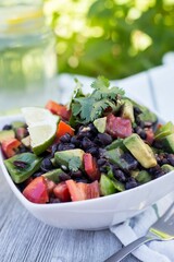 Bowl of healthy salad with lime, avocados, tomatoes, and beans with lemonade water on the gray table