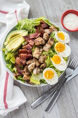 Vertical top view shot of a cobb salad on a white plate with a fork and knife near the white sauce
