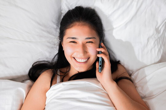 Beautiful adorable happy cheerful young asian kazachstan girl holding cellphone talking with her boyfrined on phone calling while laying in bed in the morning saying good morning. Top view.