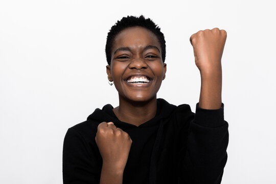 Delighted young afro american girl lady raising fists celebrating victory smiling with white teeth at camera standing over white background in studio isolated wearing casual outfit.