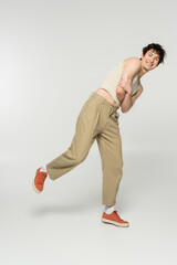 full length of cheerful nonbinary person in beige pants and sneakers posing with folded arms on grey background.