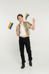 full length of positive pansexual model in black pants and snakeskin print blouse holding small lgbt flags on grey background.
