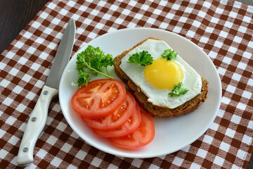 A plate of food with a toast with fried egg, parsley, tomato and a knife on a tablecloth  
