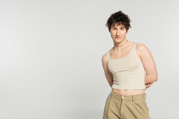 young bigender model in tank top and pants standing with hands behind back isolated on grey.
