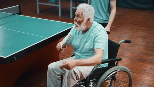 An elderly person with a disabilities in a wheelchair serves a ping pong ball while playing table tennis
