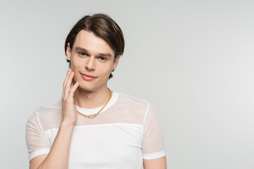 Naklejka premium positive pangender person in white t-shirt and golden necklace touching face and smiling at camera isolated on grey.