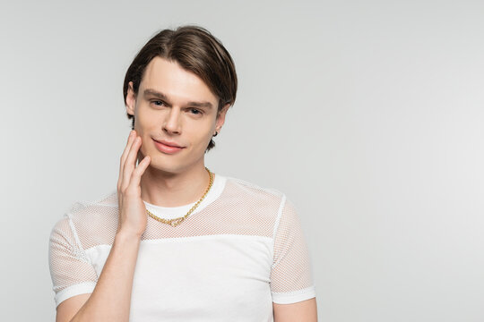 positive pangender person in white t-shirt and golden necklace touching face and smiling at camera isolated on grey.