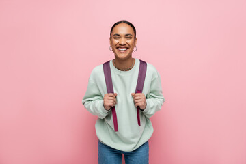Smiling african american student holding backpack isolated on pink.