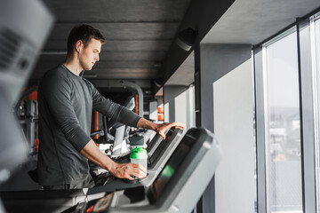 Naklejka na ściany i meble Young athletic man standing on treadmill in gym. In one hand he holds a green water bottle and the other touches the treadmill screen.