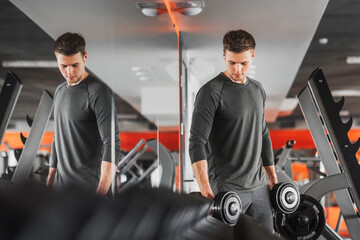 Fototapeta na wymiar Fit caucasian handsome young man in sportswear. Youth holding dumbbell during an exercise class in a gym. The athlete reflects in the mirror. Healthy sports lifestyle.