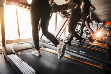 Close up photo of female legs in sportswear and sneakers running on a treadmill in the gym against the window. Sun glare from the window. Concept of healthy sports life..