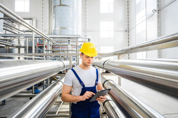 A young male specialist in a yellow hard hat uses tablet PC for controls the technological...
