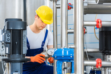 A young engineer in a yellow helmet adjusts the complex equipment of sewage treatment plants. He monitors the readings of the manometer. Steel pipes and electric motors in the foreground.