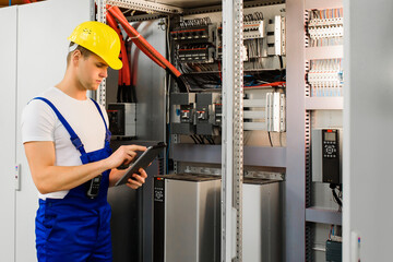 A young electrician works with a tablet standing near an industrial fuse board cabinet.