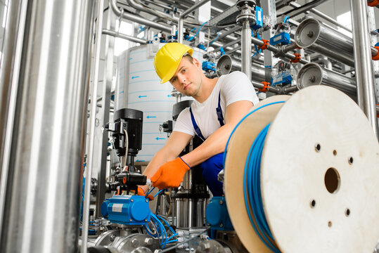 A young factory workman connects the plastic tube wire to the sensor. Industrial wastewater treatment plant on background.