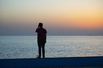 Young man watching the sea at sunset. man sitting on a wall by the ocean. Embracing the beauty and power of the ocean at sunset.