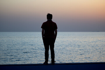 Young man watching the sea at sunset. man sitting on a wall by the ocean. Embracing the beauty and power of the ocean at sunset.