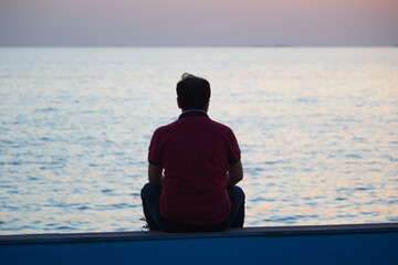 Young man watching the sea at sunset. man sitting on a wall by the ocean. Embracing the beauty and power of the ocean at sunset.