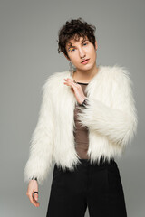 fashionable nonbinary person in white faux fur jacket looking at camera isolated on grey.