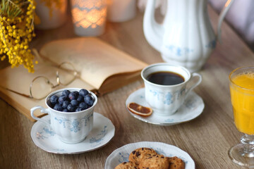 Cup of tea or coffee, plate of cookies, cup of blueberries, plate of chocolate, glass of juice, book, reading glasses, teapot, flowers and lit candles on the table. Brunch or afternoon tea concept. 