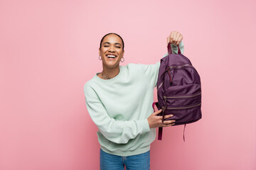 happy african american student in sweatshirt holding purple backpack isolated on pink.