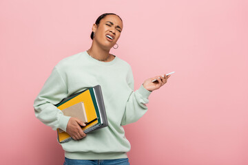 displeased african american student in sweatshirt holding gadgets and study supplies while whining isolated on pink.