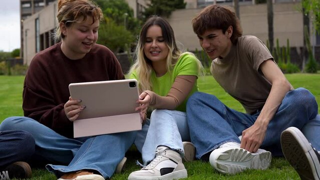 Young students having fun together taking a selfie with digital tablet while sitting outside of university meadow
