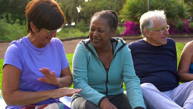 Happy senior female friends having fun together after yoga class at city park - Multiracial elderly women sitting together outdoor
