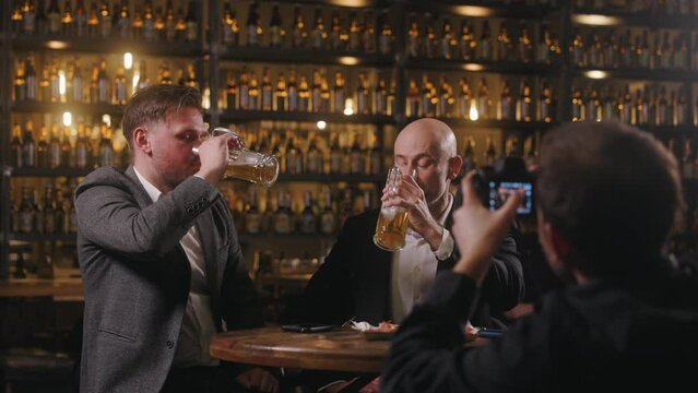 A photographer is taking a picture of two men sitting at a table drinking beer in a pub. The men are clinking glasses. The photographer is seen from behind, and the camera screen is visible.