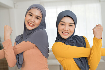 Two Asian young muslim women in hijab doing work out, stretch arm at home. Asian muslim women exercising with happy and smiling in living room - obrazy, fototapety, plakaty