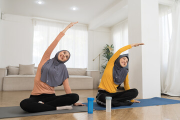 Two Asian young muslim women in hijab doing work out, stretch out arm on mat at home. Asian muslim women exercising with happy and smiling on floor in living room
