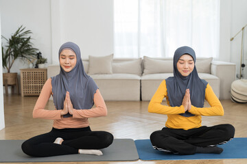 Two Asian young muslim women in hijab doing meditate yoga asana on roll mat with eyes closed in...