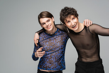 cheerful pansexual models in fashionable tops smiling and looking away isolated on grey.