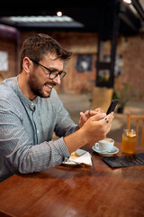 Handsome young man at cafe, sitting alone at table scrolling on his phone. Coffee break.