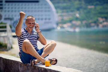 A young man is talking on the smartphone while taking a rest on the quay during a vacation on the seaside. Vacation, seaside, tourists
