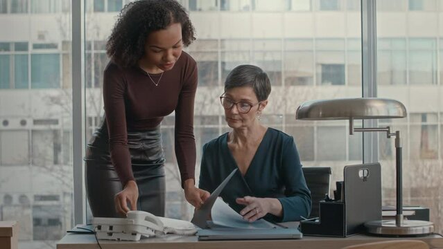 Female secretary bringing black folder with documents for sign to lady boss sitting at her desk in modern office doing paperwork
