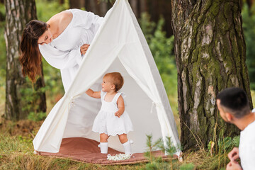 Young parents play with baby girl in toy children's wigwam house in nature. © Olha