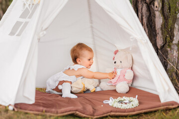 Pretty baby girl plays with teddy bear in toy outdoor wigwam.