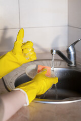 A hand in yellow rubber gloves holds a glass plate with sponge against the background of a sink with a tap and light tiles on the kitchen wall. The plot is about housework and cleanliness.