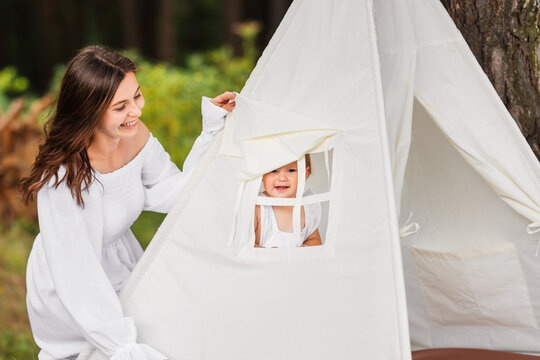 Young beautiful mother plays with her baby daughter in children's tent in nature.