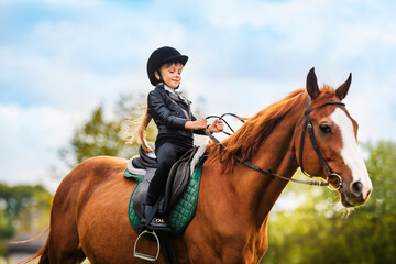 Small child in jockey outfit is riding horse on blue sky with clouds background. School of riding...