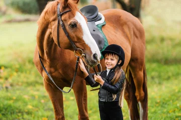 Foto op Canvas Happy child is holding horse by bridle on green meadow. Little kid in jockey outfit play with horse. © Olha