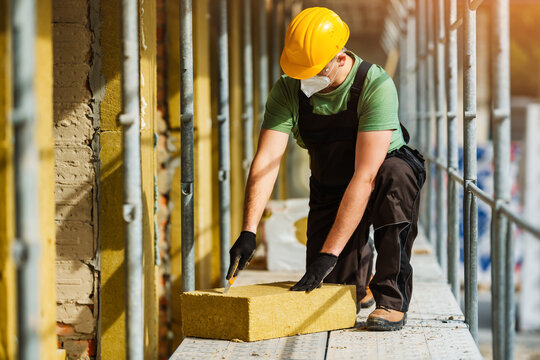 Construction worker insulates facade of house with mineral rock basalt wool mats. Builder cuts mat with knife.